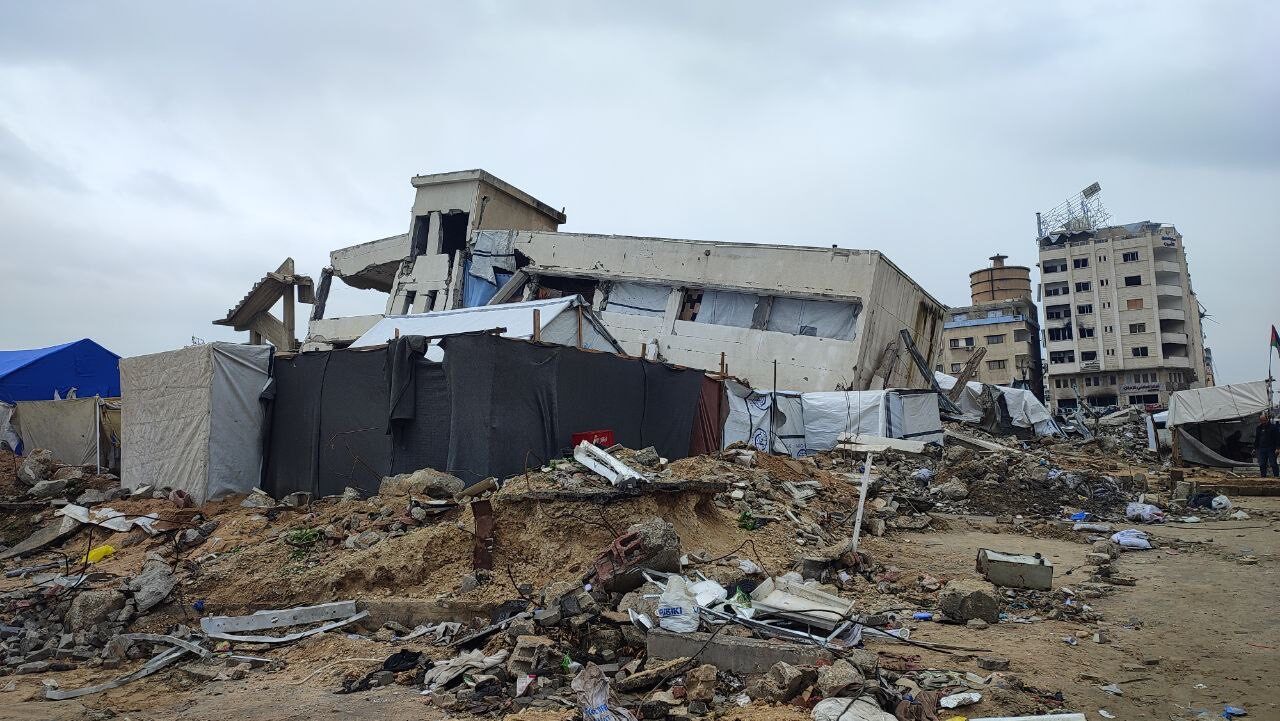 A wide street in Gaza City with every visible building along it damaged or partially collapsed, cars and debris scattered in the roadway.