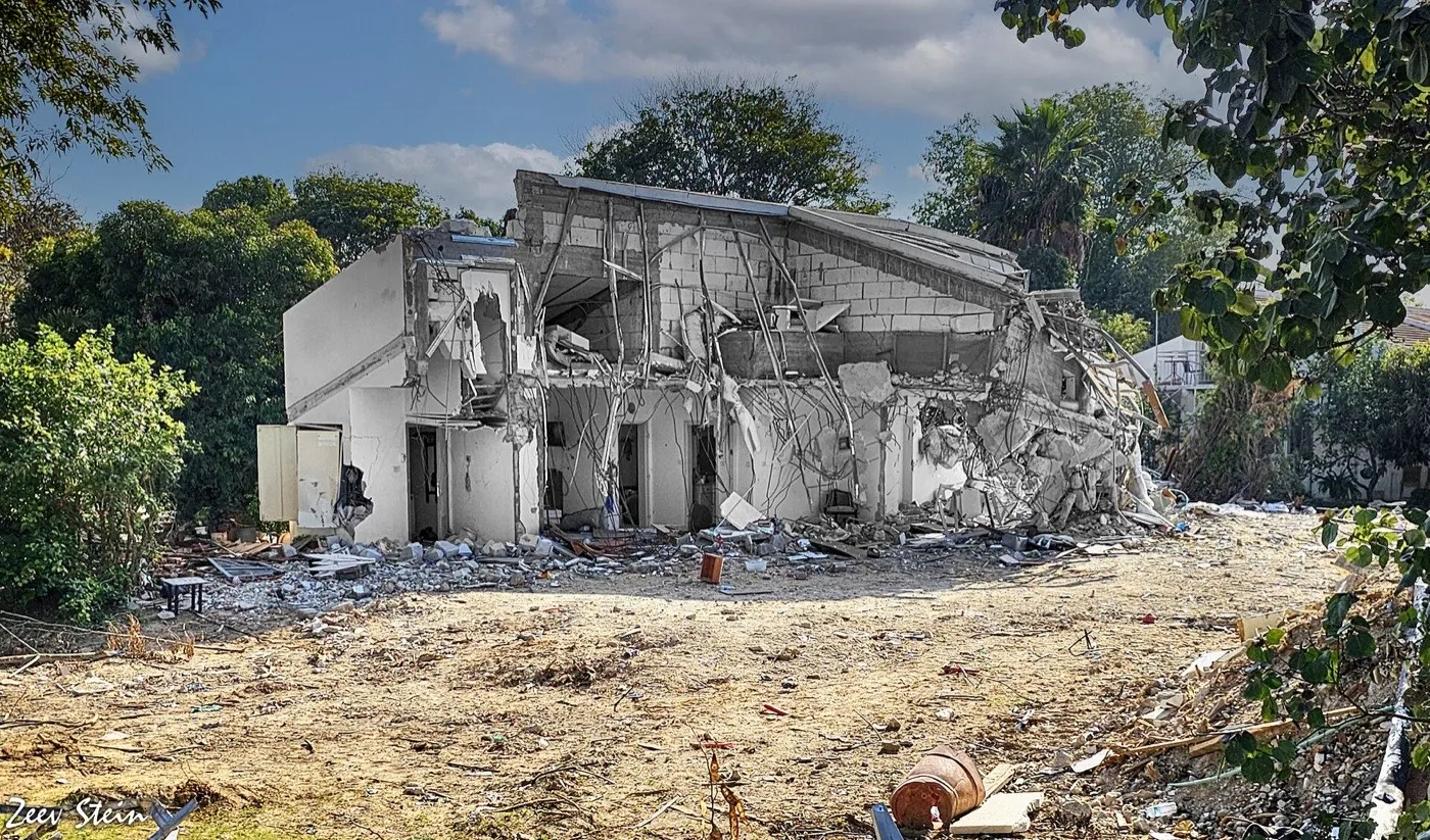 A two-story house in Kibbutz Be'eri partially collapsed into a heap of concrete, with the ground in front cleared to dirt.