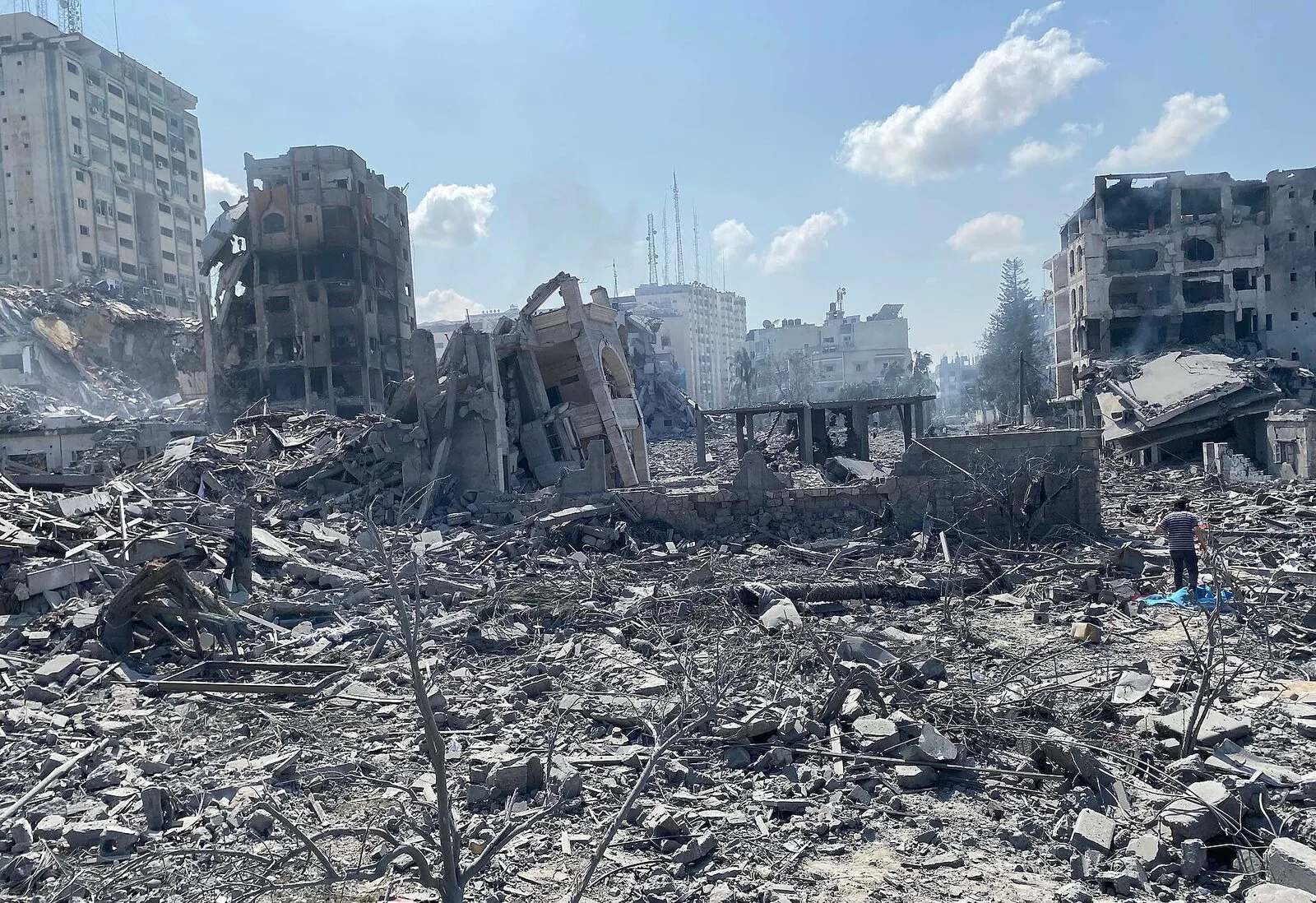 A smoke-filled street in Gaza during the early days of the war — concrete blown across the roadway, storefronts shuttered, one building leaning as if mid-collapse.