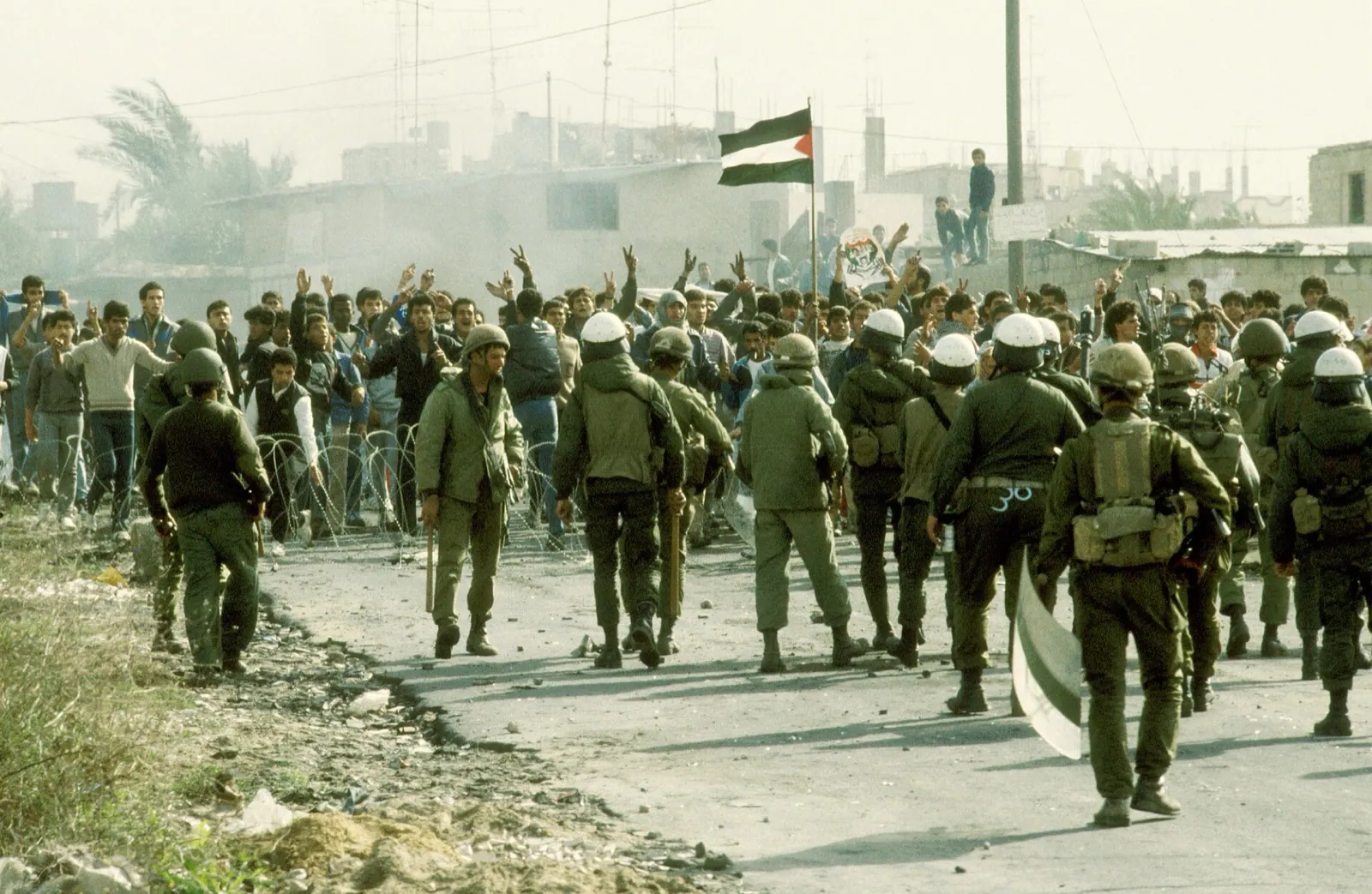Israeli soldiers in helmets face a crowd of Palestinians raising a Palestinian flag
