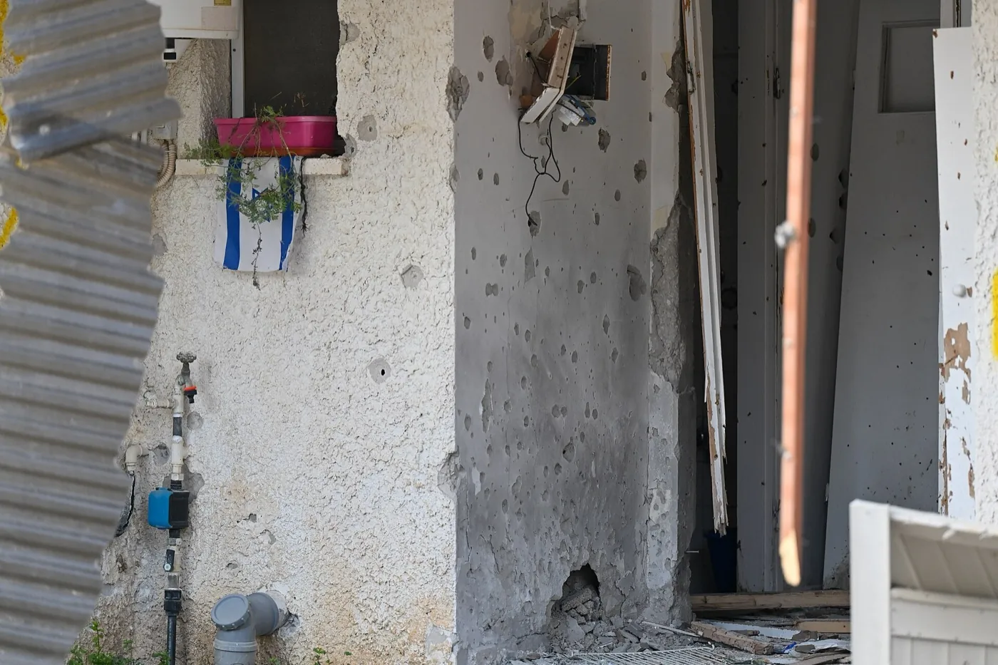 The outside wall of a house in Kibbutz Kfar Aza pocked with bullet holes, beside a doorway, with a small Israeli flag hanging from a window planter.