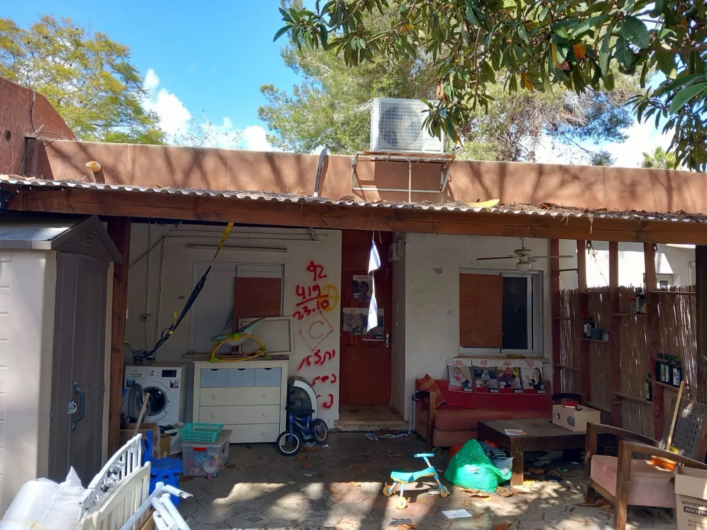 The covered porch of the Bibas family home in Nir Oz, with a washing machine, sofa, and children's ride-on toys left in place, and search markings spray-painted on the wall.